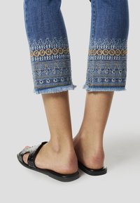 Denim capribroek met geborduurde patronen en metalen accenten aan de zoom, voorzien van een rafelrand en gecombineerd met zwarte slip-on sandalen.