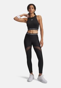 Γυναίκα ντυμένη με μαύρα αθλητικά ρούχα Under Armour με δίχτυα στα κολάν, λευκά αθλητικά παπούτσια και στεφάνι ιδρώτα στον καρπό, στέκεται μπροστά σε ένα απλό φόντο.