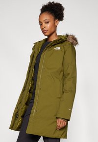 Olivově zelená zateplená parka s kapucí z umělé kožešiny, zapínáním na zip, bočními kapsami a logem The North Face na horní části levého hrudníku.