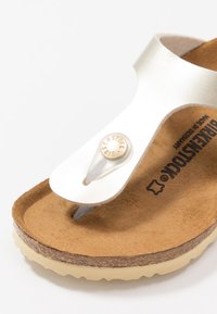 Vit syntetisk remsandal med korkfotbädd, beigefärgad gummisula och metallknappdetalj på remmen, med mjuk texturerad yta.