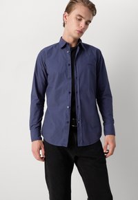 Cămașă albastru închis cu mâneci lungi, cu guler, având o textură moale și un accent subtil cu logo. Pur tată peste o cămașă neagră și pantaloni.
