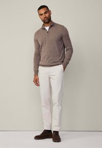 Maglione marrone con zip, pantaloni bianchi e scarpe marroni scure. Il maglione è lavorato a maglia, offrendo una finitura testurizzata. L'outfit è aderente e moderno.