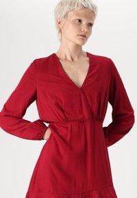 Robe enveloppante rouge avec des manches longues, décolleté en V, tissu texturé, taille cintrée et ourlet en cascade pour une dimension supplémentaire.