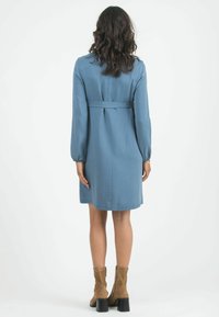 Bla dresses mit langen Ärmeln, tailliertem Schnitt und knielangem Rock, präsentiert in einer glatten Textur und minimalistischem Design, kombiniert mit hellbraunen Knöchelstiefeln.