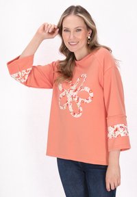 Korallfärgad långärmad topp med blommönster på ärmarna, med en blomdesign i mitten. Mjuk textur och avslappnad passform.