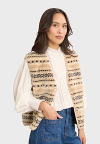 Beige Kunstfellweste mit schwarzen und braunen Mustern, ausgestattet mit einer Knopfschließe, seitlichen Taschen und einer lässigen Passform, kombiniert mit einer weißen Bluse.