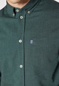 Camicia button-up di colore verde scuro realizzata in tessuto testurizzato. Presenta un colletto, bottoni visibili e una tasca con un'etichetta a strisce orizzontali.