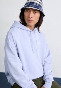 A young man with long hair wears a light purple Nike hoodie and a plaid bucket hat, seated against a plain wall with blue flooring.