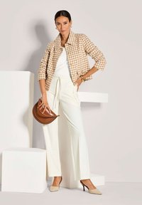Vrouw in een beige houndstooth jasje, witte top, crèmekleurige broek, beige hakken, die een bruine leren handtas vasthoudt en tegen een lichte achtergrond staat.