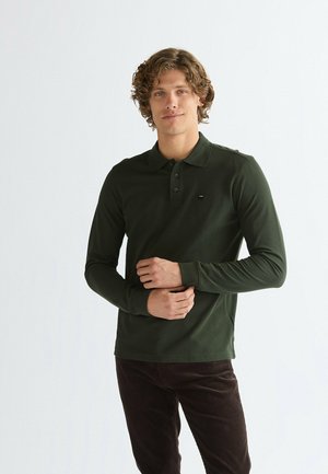 Giovane uomo con capelli ricci che indossa una polo verde scuro a maniche lunghe e pantaloni scuri, in piedi con le mani intrecciate davanti.