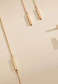 Gouden sieraden met langwerpige rechthoekige hangers aan delicate kettingen, gepresenteerd op een gestructureerde lichte achtergrond.