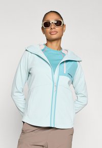 Ανοιχτό μπλε μπουφάν με φερμουάρ και κουκούλα, με ροζ λεπτομέρειες και τσέπη στο στήθος. Φορεμένο πάνω από τοπ σε τιρκουάζ χρώμα. Ομαλό, ελαστικό ύφασμα.