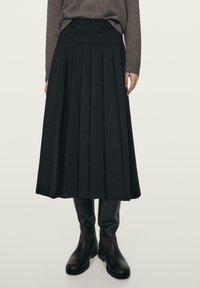 Žena v hnědém svetr, černé plisované midi sukni a černých kožených botách do poloviny lýtek stojící proti jednoduchému pozadí.