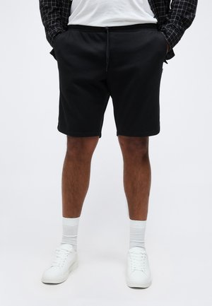 Muž oblečený v čiernych šortkách so šnúrkou, bielych ponožkách, bielych teniskách a čiernej kockovanej košeli s rukami vreckách.