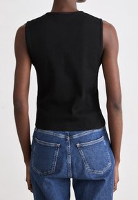 Schwarzes ärmelloses Oberteil aus Rippstoff, tailliert geschnitten. Kombiniert mit blauen Denim-Jeans mit zwei Gesäßtaschen.