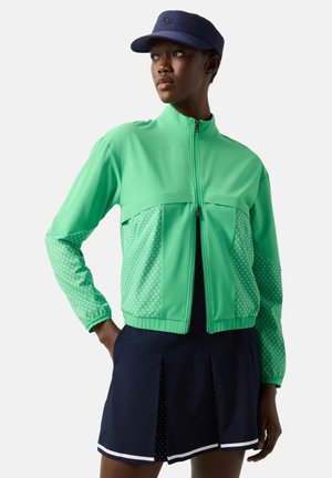 Kobieta ubrana w zieloną kurtkę na zamek z wzorzystymi rękawami, granatową spódnicę z białym wykończeniem oraz granatowy daszek, stoi z jedną ręką w kieszeni.