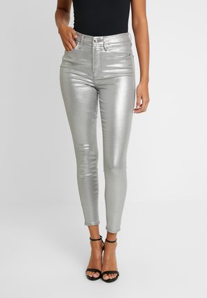 Woman wearing shiny silver high-waisted skinny jeans paired with black strappy heels and a sleeveless black top.