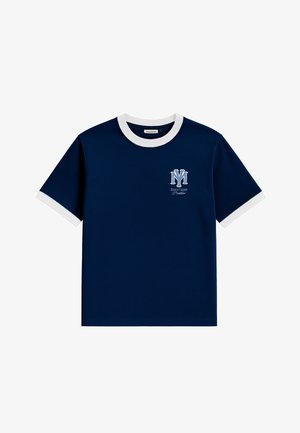 Giovane uomo che indossa un cappellino rosso e una maglietta blu scuro con bordatura bianca e logo, abbinata a pantaloni grigi a righe sottili, in piedi davanti a uno sfondo bianco.