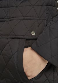 Veste matelassée noire avec un motif diagonal, poches latérales et une épingle logo dorée sur le devant. Texture douce et design élégant.