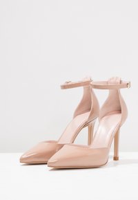 Nude lakované lodičky s vysokým podpätkom, špičatou špičkou, kotníkovým páskem a úzkým stiletto podpätkem. Hladký povrch a minimalistický design.