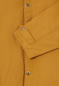 Κοντινό πλάνο μανικιού φανέλας μουστάρδιου χρώματος και κουμπωμένου μανικετόκουμπου με αντίστοιχα κουμπιά κατά μήκος του μπροστινού καπό.