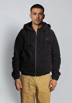 Giovane uomo di fronte, indossa una felpa nera con cerniera e coulisse, e pantaloni beige, su uno sfondo grigio chiaro uniforme.