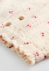 Lys beige stof med et tekstureret mønster, prydet med pink og røde blomsterbroderier samt små træknapper. Kanterne har en flæset trim.