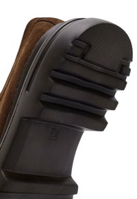 Base in camoscio marrone con suola in gomma nera testurizzata. La suola presenta scanalature evidenti e una leggera curvatura con un logo sul lato.