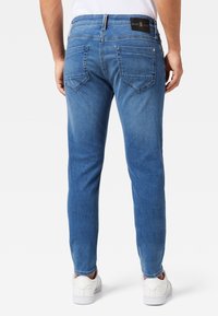 Man med medelblå slim-fit jeans och vita sneakers, stående med ryggen mot kameran mot en vit bakgrund.