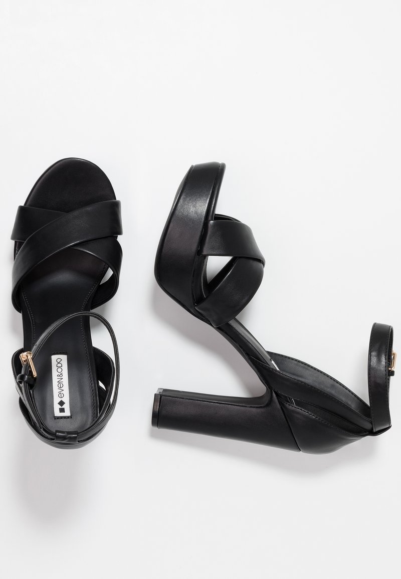 Sandalias negras de tacón alto con tiras cruzadas en cuero suave, que cuentan con un tacón cuadrado y una correa en el tobillo con herrajes en tono dorado.