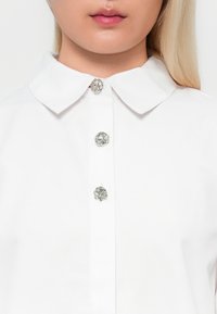 Vit blus med krage som har fem dekorativa silverknappar framtill. Slät tyg med en krispig textur och rundad krage.