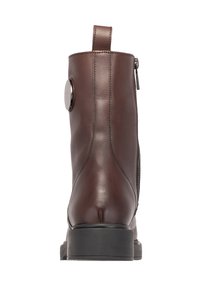 Bottine en cuir marron avec une finition lisse, fermeture éclair à l'arrière et accent en métal circulaire. Dispose d'une semelle noire basse et robuste.