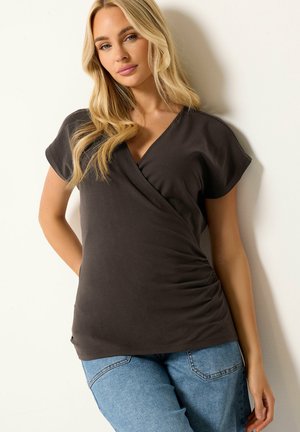 Blonde vrouw die een donkerbruine omslagstijl korte mouwen top en lichtblauwe spijkerbroek draagt, staand tegen een witte muur met één hand achter haar rug.