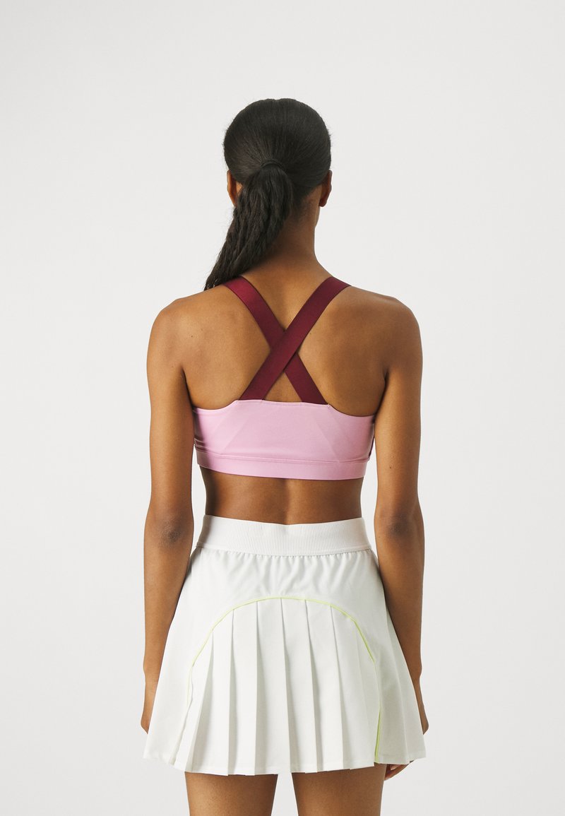 Sujetador deportivo en rosa con tirantes cruzados color marrón, combinado con una falda blanca con pliegues que presenta acentos en verde neón a lo largo de las costuras.