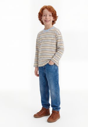 Glunderende roodharige jongen staand met één hand in zijn zak, een gestreept shirt met lange mouwen, blauwe spijkerbroek en bruine schoenen op een witte achtergrond.