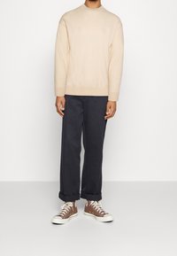 Suéter de punto beige con cuello redondo, combinado con pantalones oscuros de tiro alto y zapatillas altas marrones con suelas y cordones blancos.