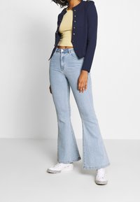 Marinblå croppad jacka med silverknappar, randig gul topp, ljusblå vida jeans och vita sneakers med rutigt mönster.