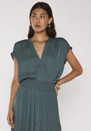 Donna con lunghi capelli scuri che indossa un vestito verde acqua con scollo a V e orecchini a cerchio dorati, posando con una mano sulla vita su uno sfondo semplice.