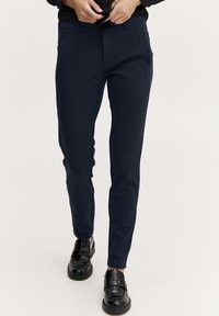 Námořnické modré slim-fit kalhoty s předními kapsami, poutky na pásek a hladkým povrchem, kombinované s černými mokasíny. Minimalistický design, žádné viditelné vzory.