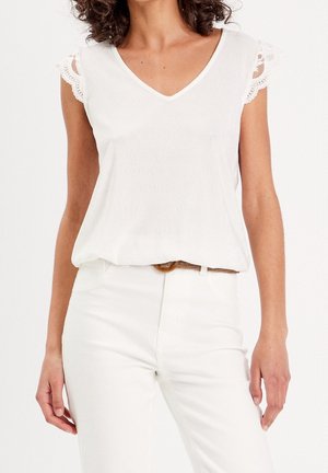 Femme portant une blouse blanche à col en V avec des manches courtes en dentelle et un pantalon blanc, debout devant un fond clair uni.