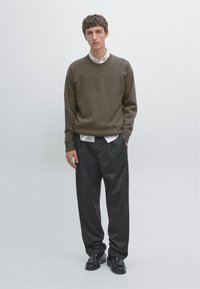 Bruine gebreide trui met geribbelde manchetten, gedragen over een witte blouse met kraag. Zwarte wijde broek en zwarte instappers maken de look af.