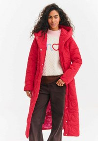 Czerwona pikowana kurtka puchowa z kapturem, z fakturowanym wzorem i przednimi guzikami, noszona na jasnym swetrze i brązowych spodniach.