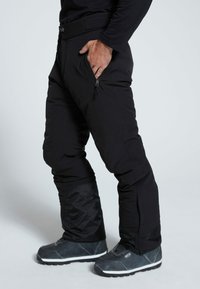 Schwarze isolierte Hose mit Reißverschlusstaschen, hoher Taille, schmal zulaufenden Beinen und verstärkten unteren Säumen, kombiniert mit grauen Stiefeln.