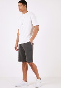 Hvid T-shirt med korte ærmer parret med mørkegrå shorts. Shortsene har sidelommer, og outfittet suppleres af hvide sneakers.