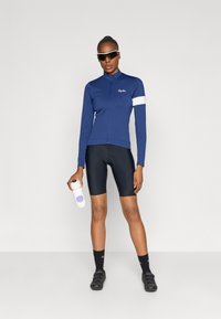 Camisola de ciclismo de manga longa azul-marinho com risca branca, calções de ciclismo pretos, sapatos pretos, segurando uma garrafa de água branca com acento roxo.