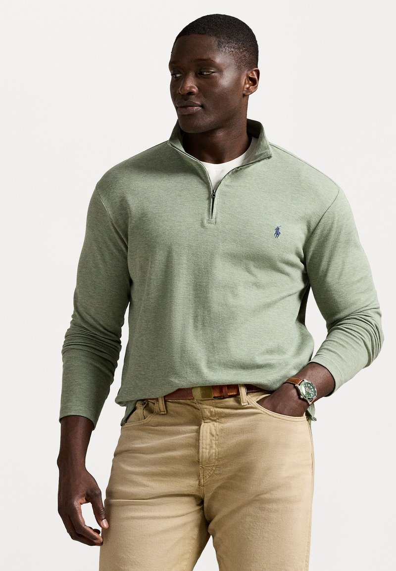Light green long-sleeve pullover with a zippered collar and a small blue logo. Worn with khaki pants and a brown belt. Textured fabric.