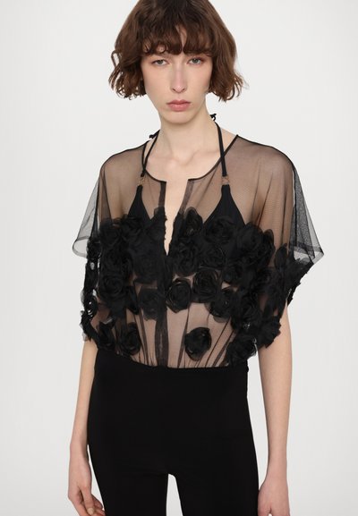 Schwarze durchsichtige Bluse mit schwarzen Blumenapplikationen, kurzärmlig und mit einem Schlüssel-Ausschnitt. Kombiniert mit schwarzen Leggings.