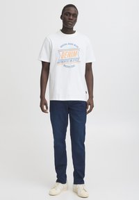 Valge puuvillane t-särk sinise ja oranži graafilise tekstiga, kombineeritud tumeliste teksade ja valgete spordijalatsitega, millel on beežid aktsendid.