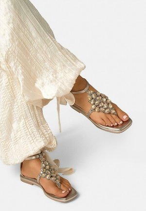 Pies que llevan sandalias plateadas metálicas con grandes adornos de cristal, combinadas con pantalones blancos fluidos y texturizados.