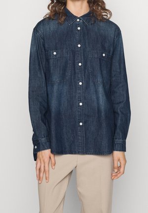 Osoba nosící tmavě modrou džínovou košili s knoflíky a kapsami na hrudi, béžové kalhoty a viditelné dlouhé kudrnaté hnědé vlasy.